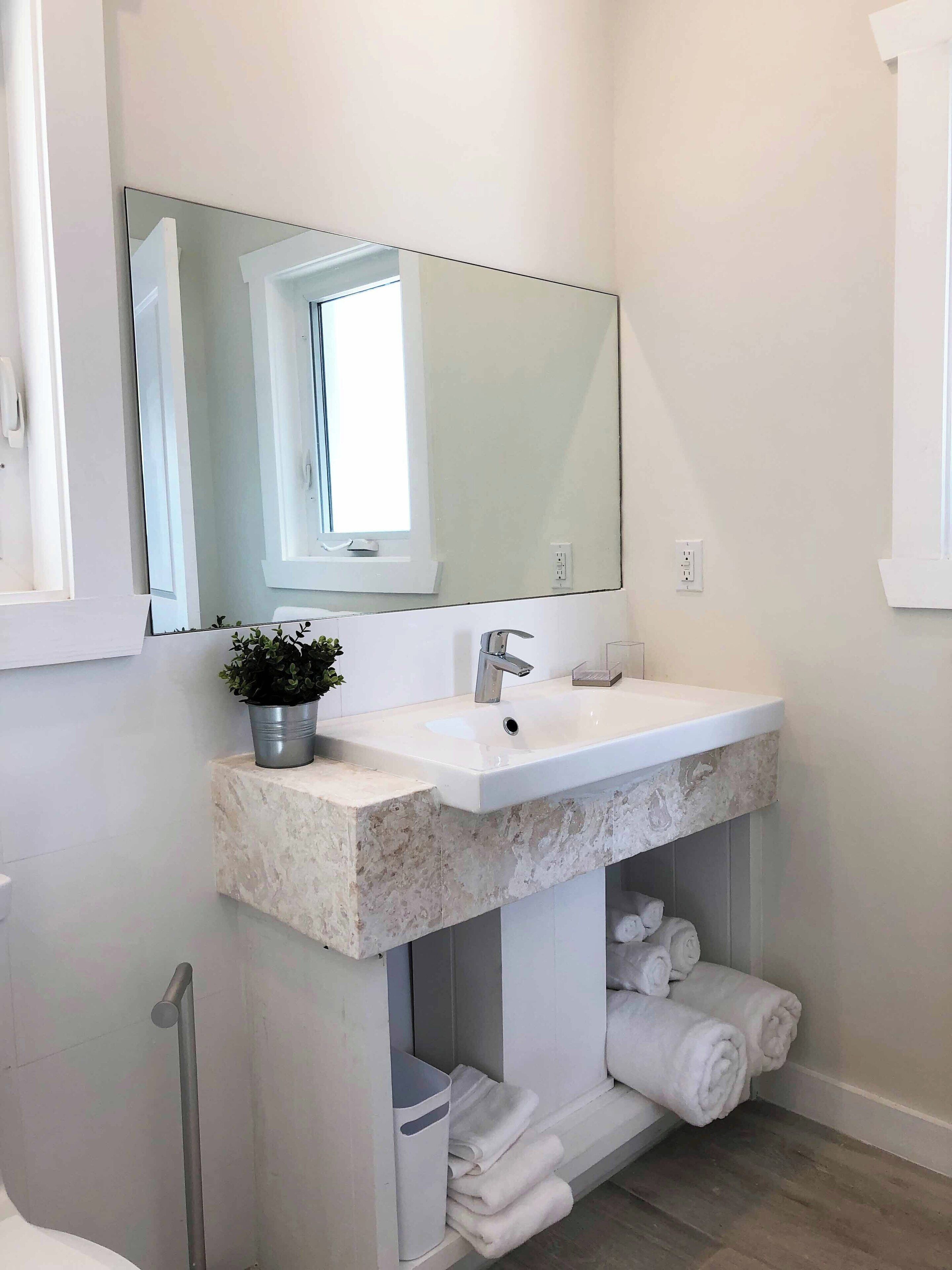 A modern bathroom with a large mirror, white sink, and marble countertop. There is a potted plant on the countertop and towels hanging on the wall.