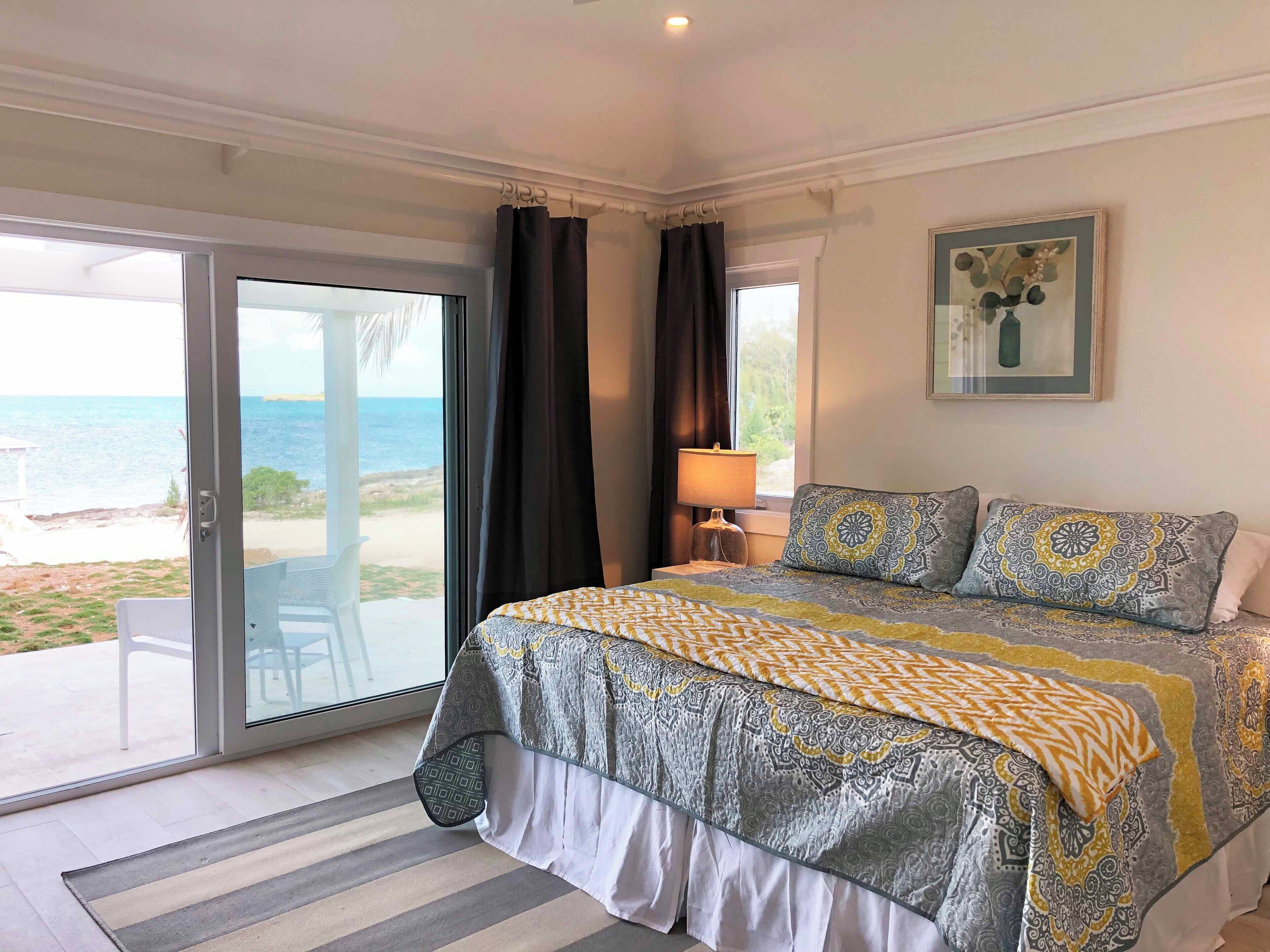 Quarto com cama, janela com cortinas, luminária e vista para a praia.
