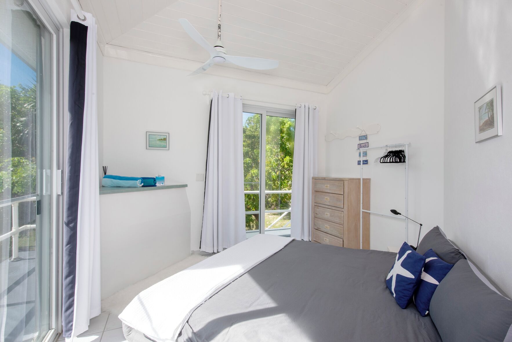 Ein Schlafzimmer mit Bett, Kommode, Fenster mit Vorhängen, Deckenventilator und Blick ins Grüne.