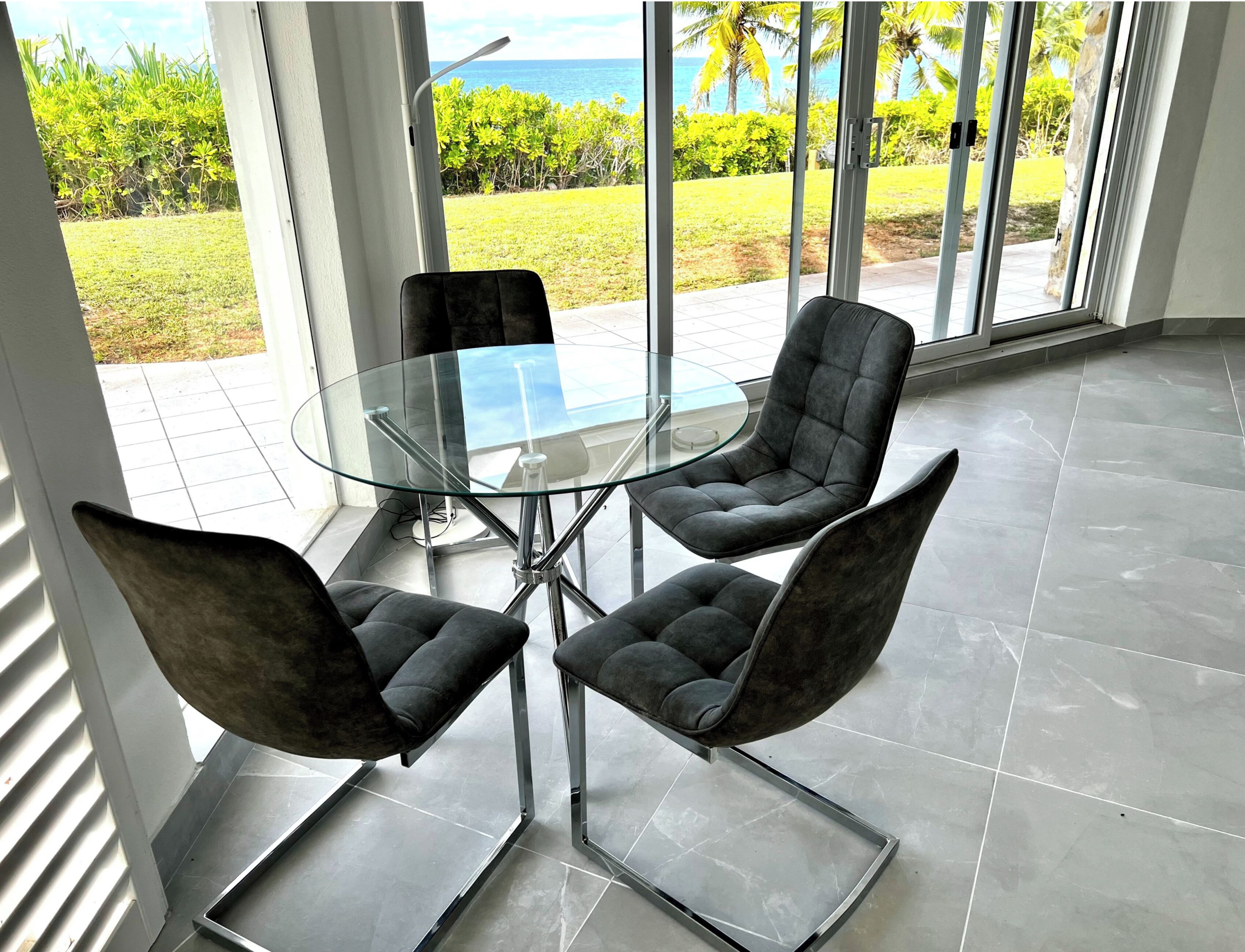 A modern dining area with a glass table and four upholstered chairs, large windows with a view of the ocean and greenery.