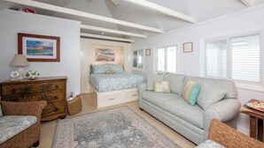 A bedroom with a bed, a grey sofa, a wooden dresser, a patterned rug, and a ceiling fan. - Harbour Island Haven: Newly Remodeled Cottage Near Pink Sands Beach (Dunmore Town)