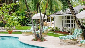 Un área junto a la piscina con sillones de mimbre, un pequeño pabellón y una palmera.