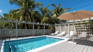 A swimming pool with a wooden deck, white lounge chairs, and a pergola with an umbrella. - Pink Sand Paradise: Cozy Beachfront Cottage (Governor's Harbour)
