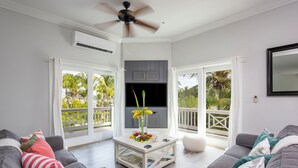 A spacious living room with grey sofas, a coffee table with flowers, a ceiling fan, and a TV. - Pink Sand Paradise: Cozy Beachfront Cottage (Governor's Harbour)