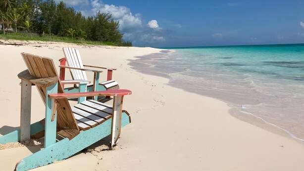 A beach with two wooden Adirondack chairs and a table, a clear blue sky, and a calm sea. - Pink Sands Retreat: Beachfront Cottage With Views (North Palmetto Point)