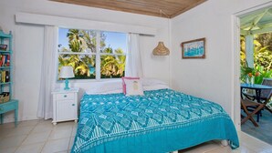 A bedroom with a bed, a nightstand, a window with curtains, a bookshelf, and a view of palm trees. - Pink Sands Retreat: Beachfront Cottage With Views (North Palmetto Point)
