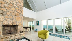 A spacious living area with a stone fireplace, a dining table, and a large window overlooking the ocean. - Island Escape: Secluded Beachfront Cottage With Pool (North Palmetto Point)