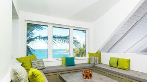 A modern living room with a large window overlooking the ocean, a built-in bench with cushions, and a coffee table. - Island Escape: Secluded Beachfront Cottage With Pool (North Palmetto Point)