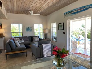 A living room with a sectional sofa, a glass coffee table, and a vase of flowers. There are windows with views of greenery and a door leading outside.