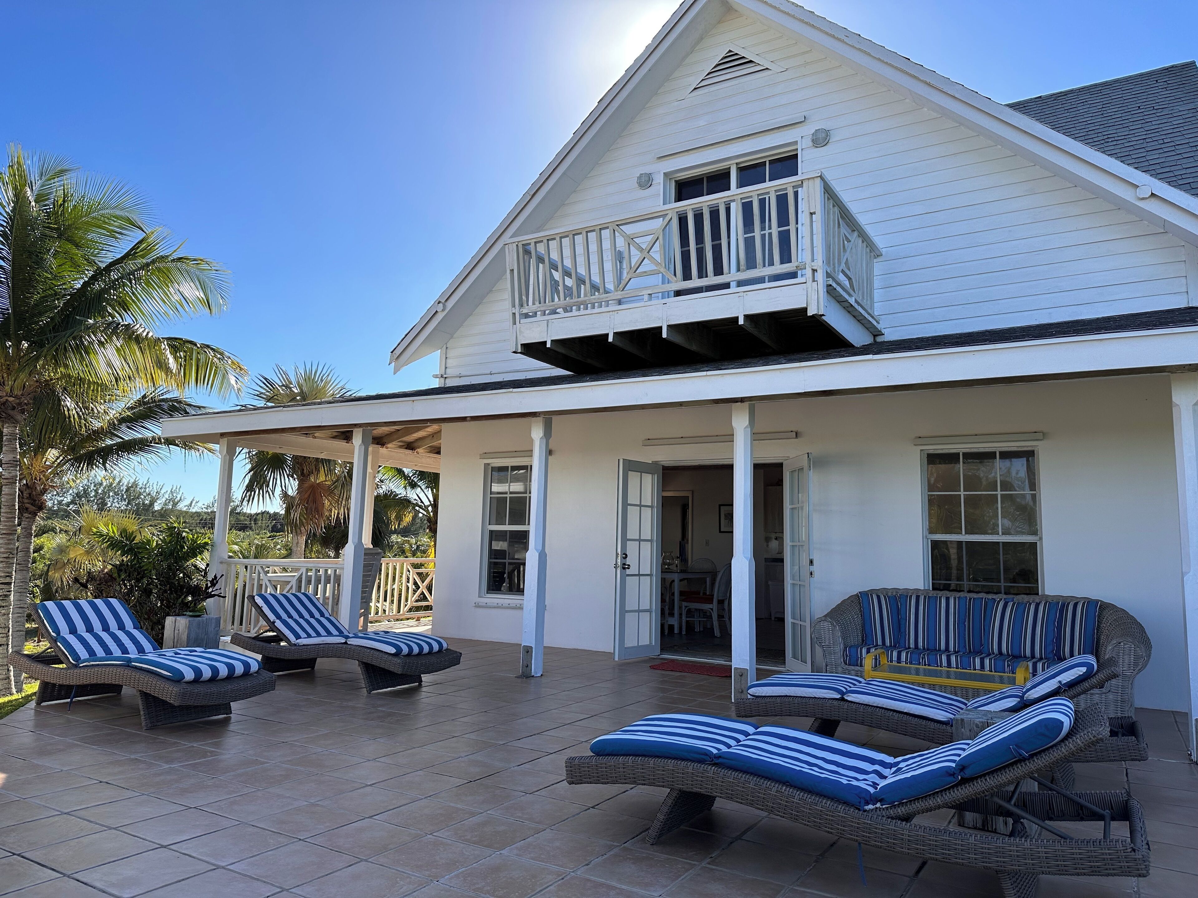 Une maison blanche avec un patio couvert, deux chaises longues dotées de coussins à rayures et un petit balcon.