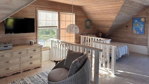 Une chambre spacieuse située sous les combles, avec un plafond en bois, une grande fenêtre, un téléviseur à écran plat, un fauteuil en osier, une commode et une carte accrochée au mur.