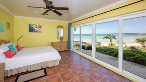 A bedroom with a bed, a dresser, a ceiling fan, and large windows offering a beach view. - Banks Road Bliss: Luxury Estate With Pool (South Palmetto Point)