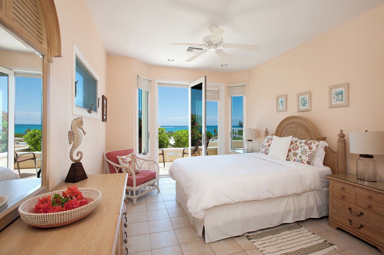 A bedroom with a bed, a chair, a bedside table, a lamp, and a view of the ocean.