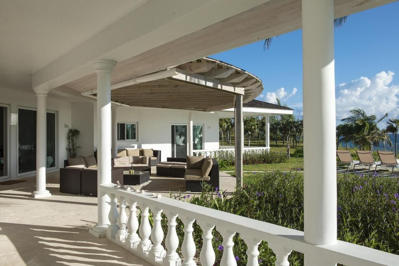A covered outdoor seating area with white columns and a wooden ceiling, overlooking a landscaped garden and pool area.
