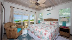 Un dormitorio con una cama grande, una silla de mimbre, vista al océano, ventilador de techo y una ventana con persianas.