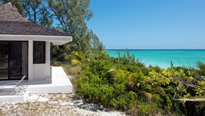 A beachside property with a white building, a patio with a metal chair, and a view of the turquoise sea. - Tropical Hideaway: Charming Cottage Near Pink Sands Beach (Rock Sound)