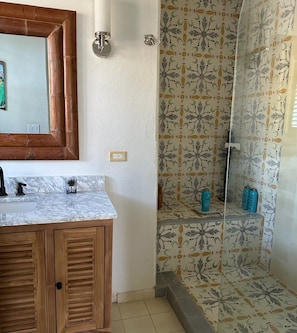 A bathroom with a wooden vanity, a mirror, a shower with patterned tiles, and a shelf with toiletries. - Shangri-La Serenity: Beach Villa With Views (Governor's Harbour)