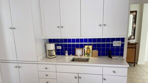 Une cuisine avec des meubles blancs, une machine à café, un évier et un plan de travail bleu recouvert de carreaux.