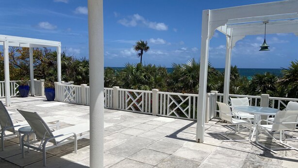Une terrasse extérieure couverte, avec des chaises et une table blanches, des rampes blanches et une vue sur l’océan.