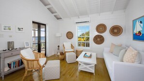 Ein heller, weißer Innenraum mit Holzboden, Korbmöbeln, einem weißen Sofa und Blick durch eine Glastür.
