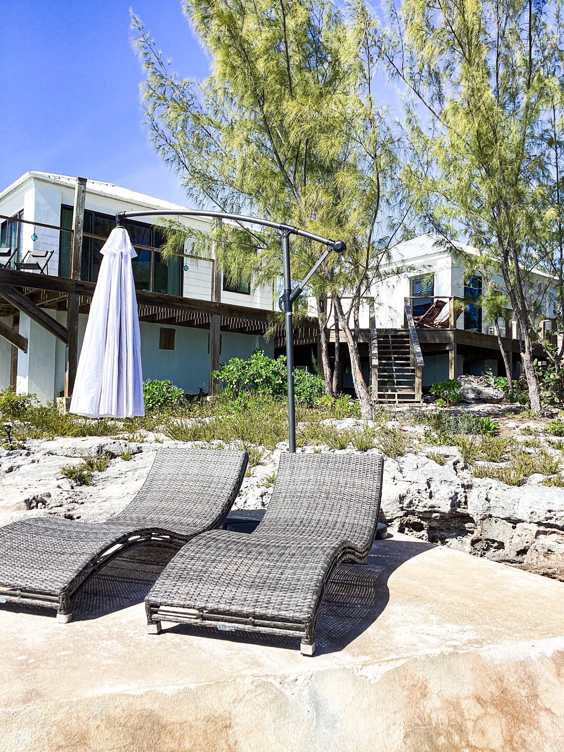A beachside property with two lounge chairs, a covered umbrella, and a modern building with a balcony.