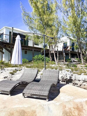 A beachside property with two lounge chairs, a covered umbrella, and a modern building with a balcony. - Seaside Serenity: Elegant Beachfront Escape (Rainbow Bay)