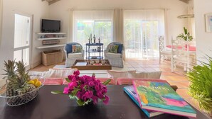Amplia sala de estar con techo alto, televisor en un estante, mobiliario de mimbre, una mesa de centro con un jarrón de flores y vistas al exterior a través de amplias ventanas.