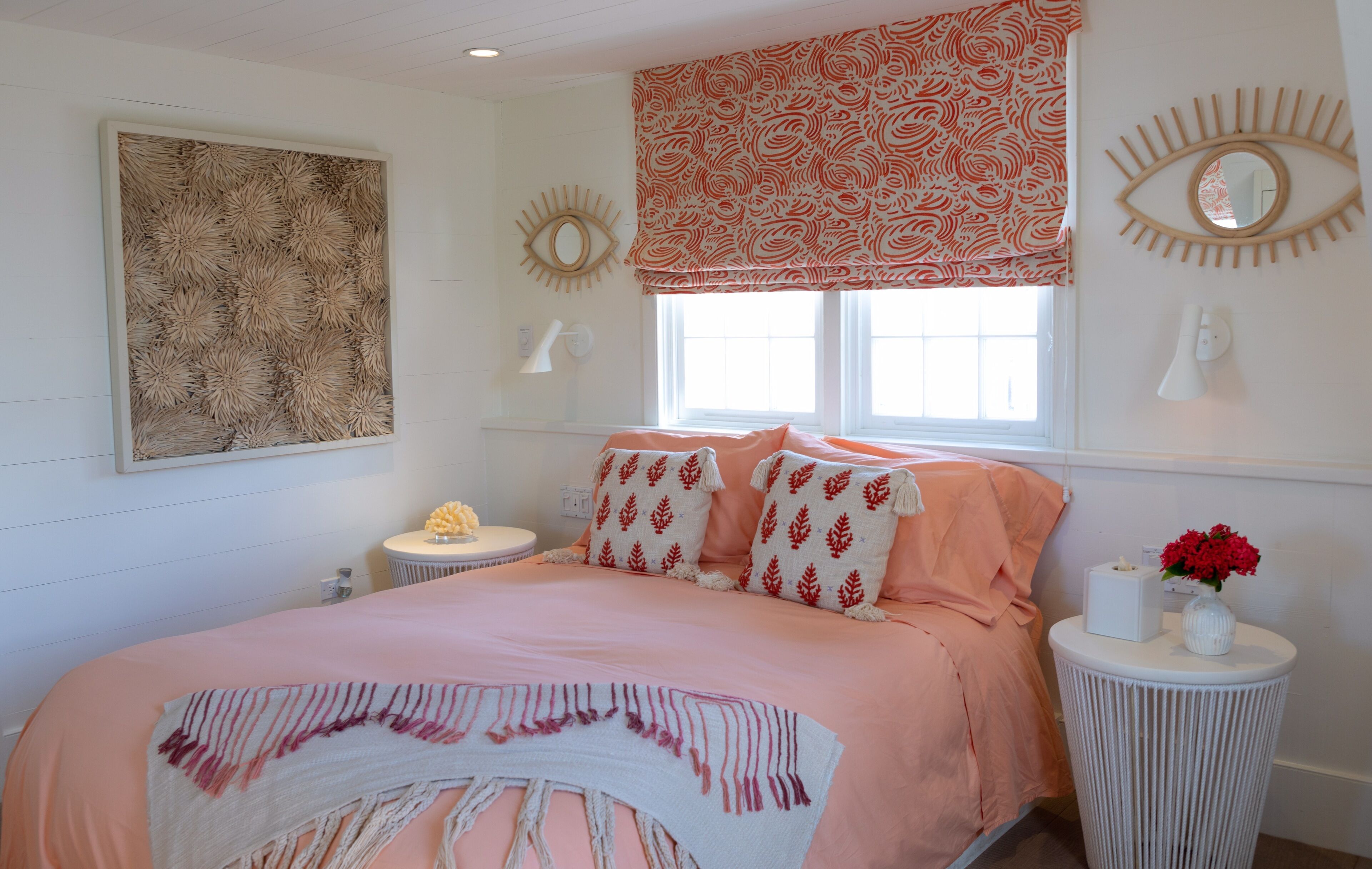 A bedroom with a large bed, a round side table, a window with patterned curtains, and a wall-mounted eye decoration.