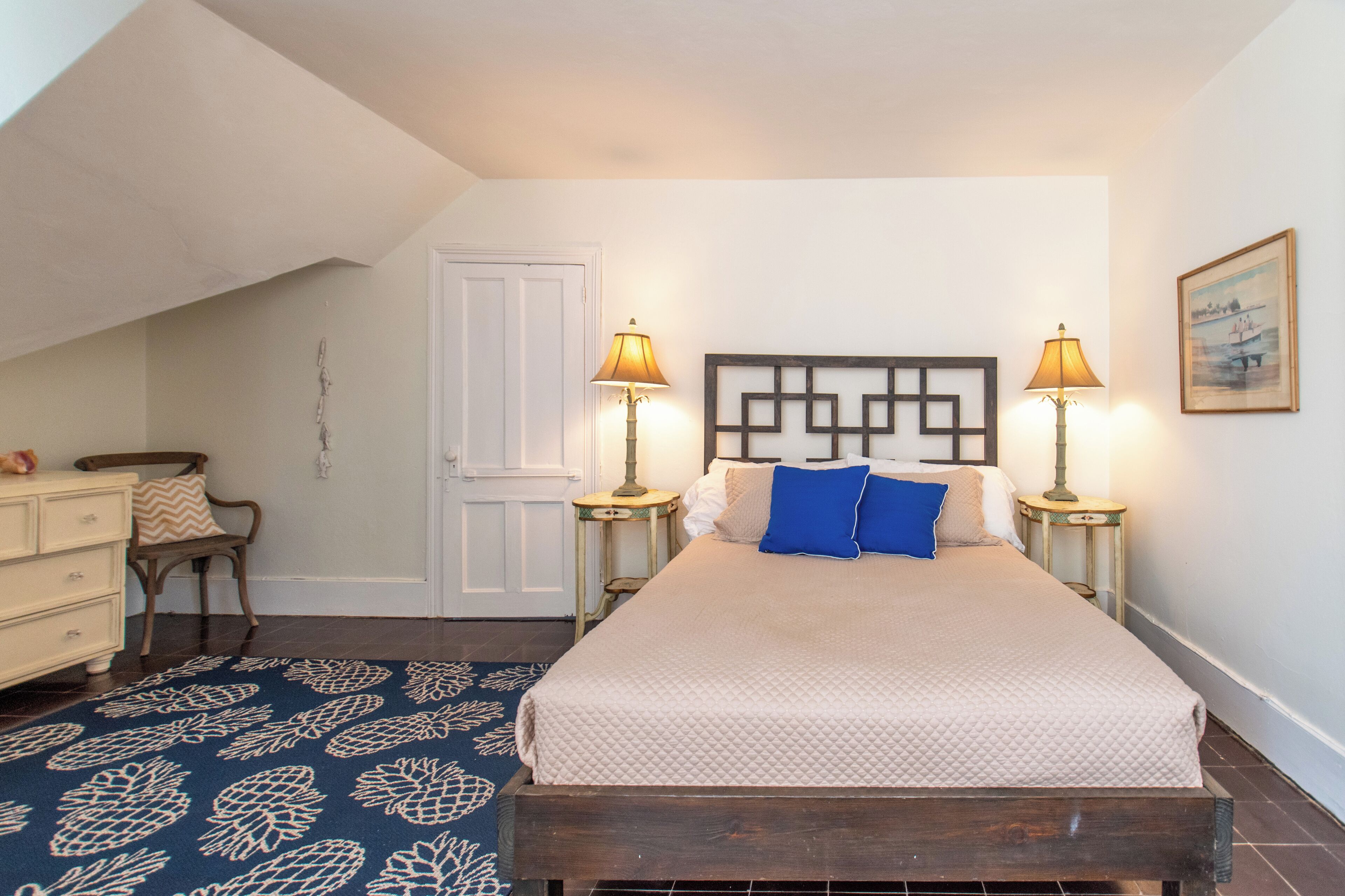 A bedroom with a bed, two bedside lamps, a dresser, a chair, a patterned rug, and a door.