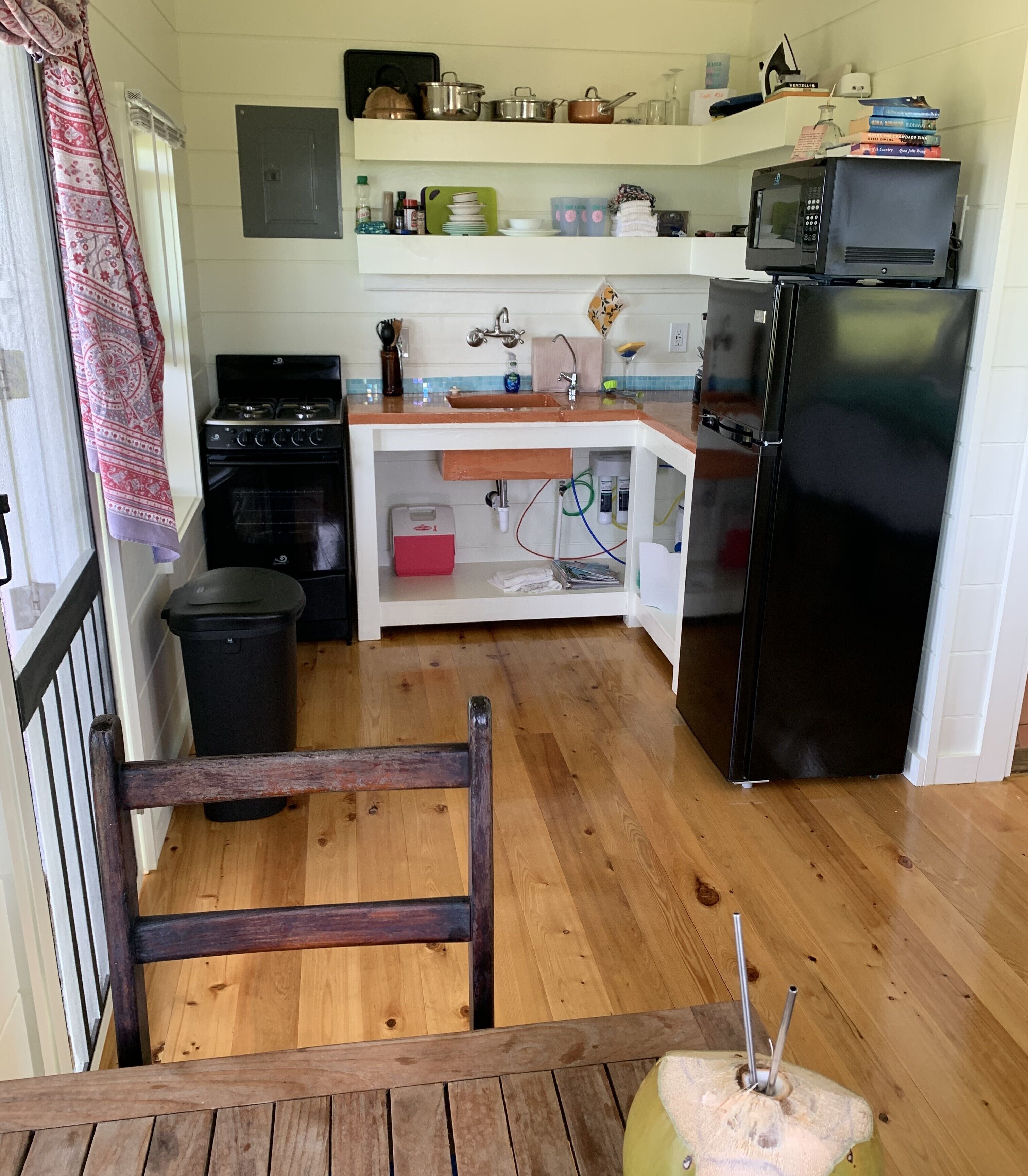 A compact kitchen with a stove, sink, and refrigerator. There is a wooden chair in the foreground and a trash bin to the left.