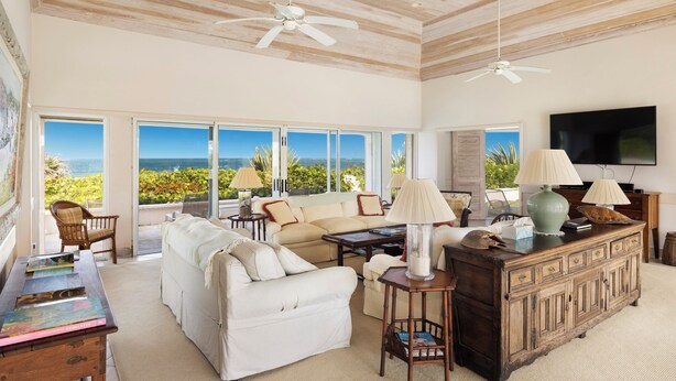 A spacious living room with a large window overlooking a scenic view, a ceiling fan, and a wooden cabinet with lamps. - Windermere Paradise: Luxurious Estate With Amenities (Governor's Harbour)