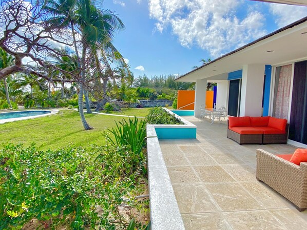 A modern outdoor patio with a pool, a dining area, and a seating area with a red sofa. - Coastal Haven: Newly Renovated Beachfront Cottage With Pool (Governor's Harbour)