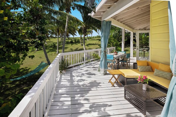 A wooden deck with a white railing, a yellow sofa, and a table with a vase of flowers. - Poponi Cottage Paradise: Pink Sand Retreat (North Palmetto Point)