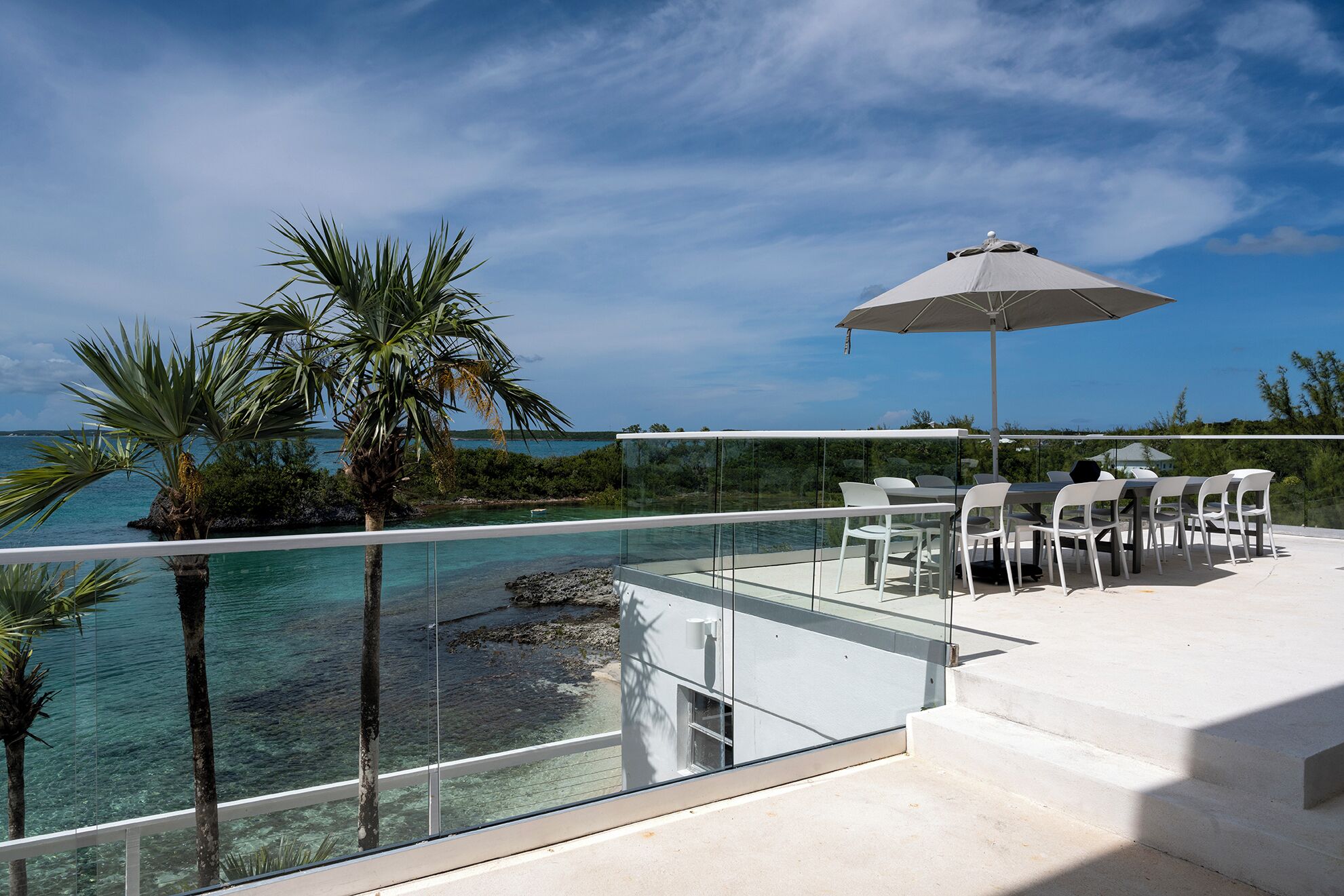 Un espacio al aire libre con paredes de vidrio, una piscina, sillas blancas y una mesa bajo un paraguas grande.
