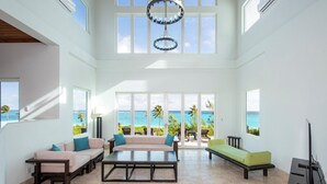 A spacious living room with a large window overlooking the ocean, a ceiling fan, and a flat-screen TV. - Contemporary Comfort: Stunning Villa With Pool & Restaurant (Governor's Harbour)
