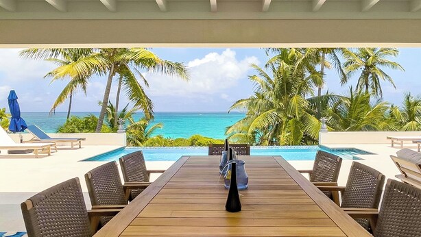 A wooden table with chairs and a vase, overlooking a pool with palm trees and the ocean. - Pink Sand Paradise: Luxury Home With Heated Pool (North Palmetto Point)