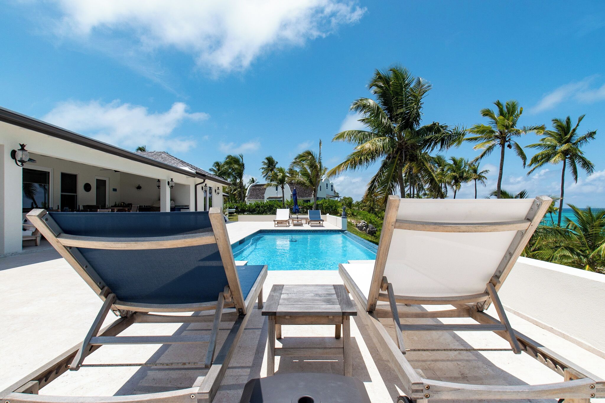 Un espace aménagé au bord de la piscine, avec deux chaises longues et une table, d’où l’on peut admirer la piscine et les palmiers.