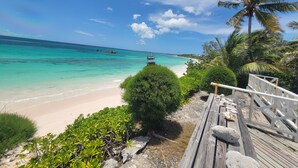Una terraza de madera que da a una playa con agua turquesa cristalina, un barco a lo lejos y palmeras.