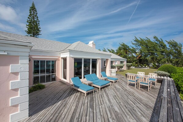 A covered outdoor deck with lounge chairs and a table, surrounded by a wooden deck and a building with large windows.