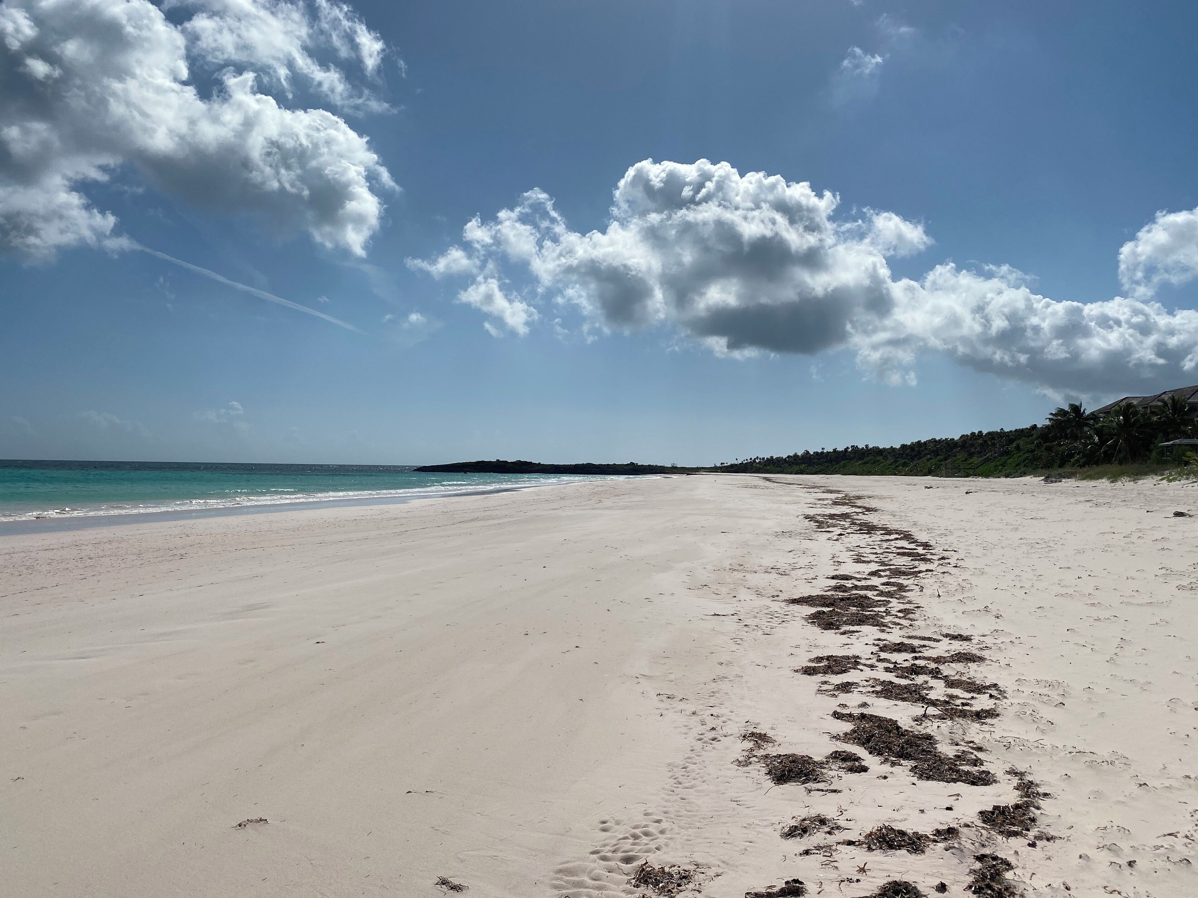A wide sandy beach with footprints leading to the water, a clear sky, and a distant hill with vegetation.