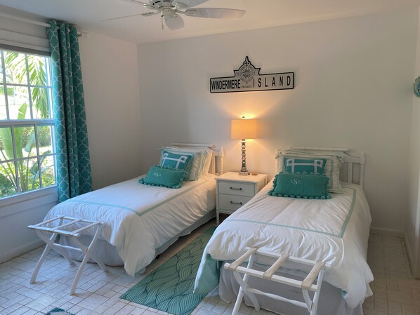 A bedroom with two beds, a ceiling fan, a nightstand with a lamp, and a sign on the wall. - Coastal Chic: Stylish Condo On Windermere Beach (Governor's Harbour)