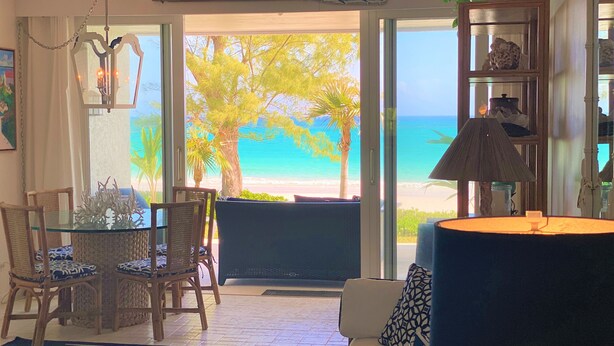 A room with a glass table, wicker chairs, a sofa, and a view of the beach. - Coastal Chic: Stylish Condo On Windermere Beach (Governor's Harbour)