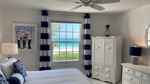 A bedroom with a bed, a ceiling fan, a window with a view of the beach, a white cabinet, and a lamp. - Coastal Chic: Stylish Condo On Windermere Beach (Governor's Harbour)