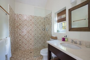 A bathroom with patterned tiles, a glass shower enclosure, a toilet, a sink with a mirror, and a window with a view of greenery.