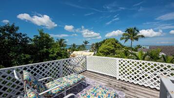 A wooden deck with white lattice railings, two lounge chairs with colorful cushions, and a view of the ocean and palm trees.