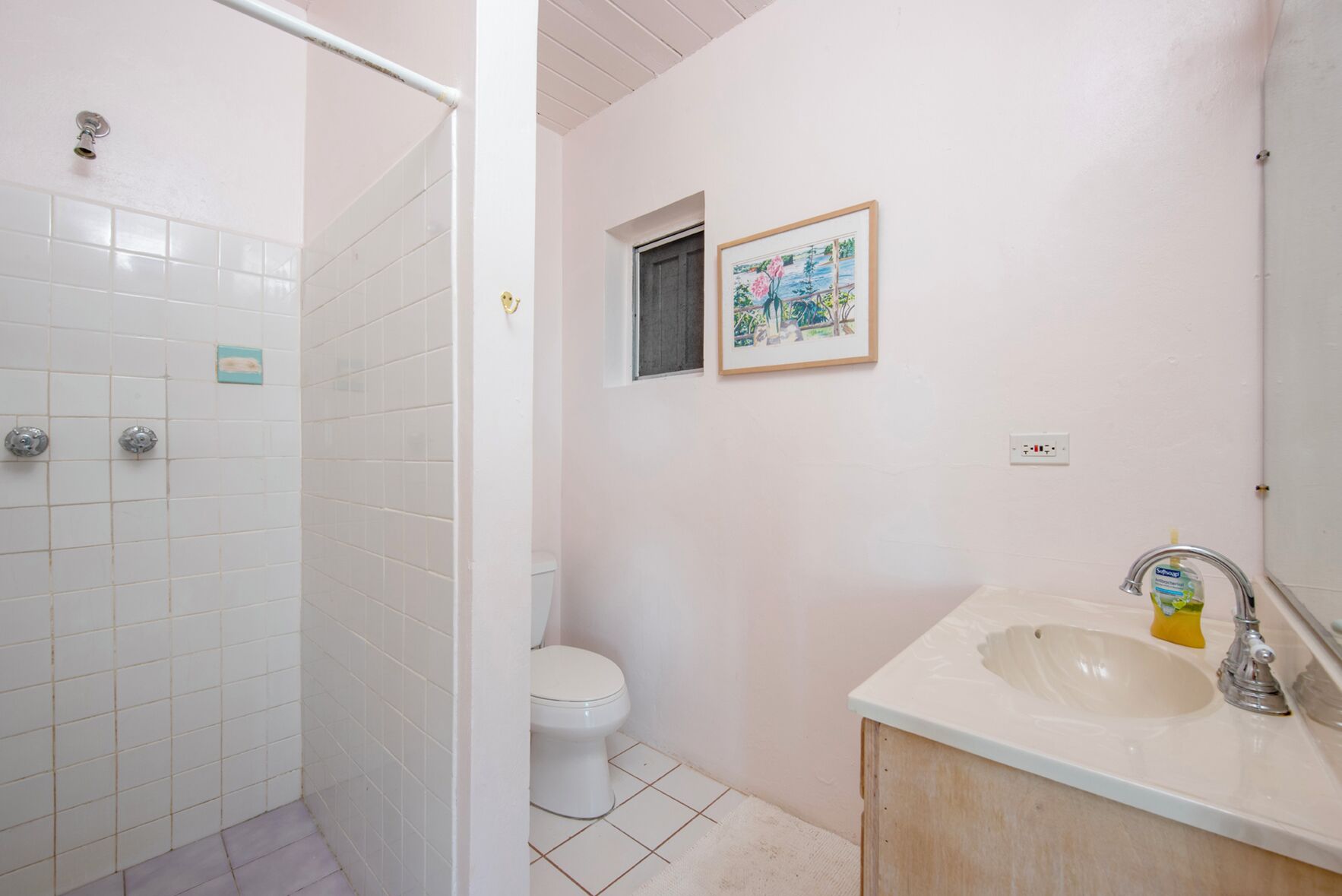 Une salle de bain avec des carreaux blancs, des toilettes, un lavabo avec un miroir et un tableau encadré au mur.