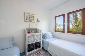 Quarto pequeno e bem iluminado com uma cama de solteiro, uma mesinha com abajur e uma janela com vista para árvores.
