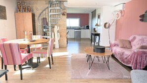 Een moderne woonkamer met een roze bank, een houten eettafel met gestreepte stoelen, een spiltrap en een keuken op de achtergrond.