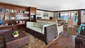 A cozy living room with a large wicker sofa, a bookshelf filled with books, and a view of the sea through large windows. - Beachfront Bliss: Private Retreat With Spectacular Views (Blowing Point)