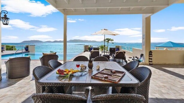 A covered outdoor dining area with wicker furniture, a table set with fruits, wine, and a vase, overlooking a pool and the sea. - Beachfront Bliss: Private Retreat With Spectacular Views (Blowing Point)